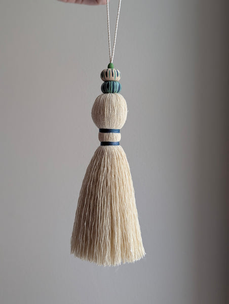 A close-up of a handmade wooden bead tassel, featuring a cream-colored cotton and blue and green accents against a soft beige background.