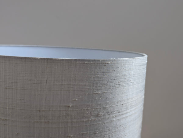 Close-up of a drum lampshade made of textured silk in a light warm gray or off-white color, showing the inner lining and fine horizontal weave details.