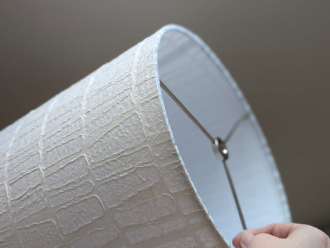 A close-up of a white textured lampshade with a nickel spider washer, with a hand visible at the top right.
