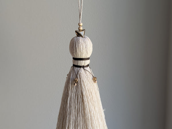 A close-up of a decorative tassel with cream-colored thread, a brown wrapped detail, and brass and wooden beads shown against a neutral gray background