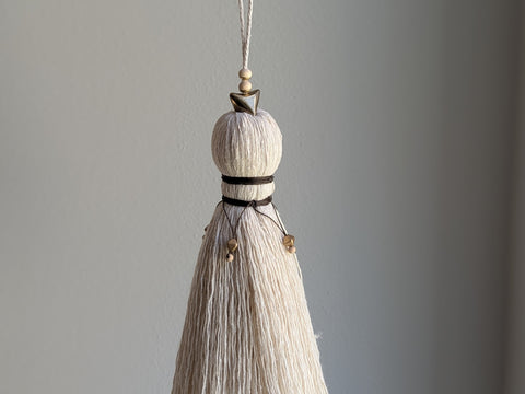 A close-up of a decorative tassel with cream-colored thread, a brown wrapped detail, and brass and wooden beads shown against a neutral gray background