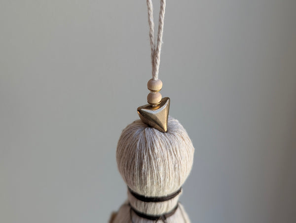 A close-up of a decorative tassel with cream-colored thread, a brown wrapped detail, and brass and wooden beads shown against a neutral gray background