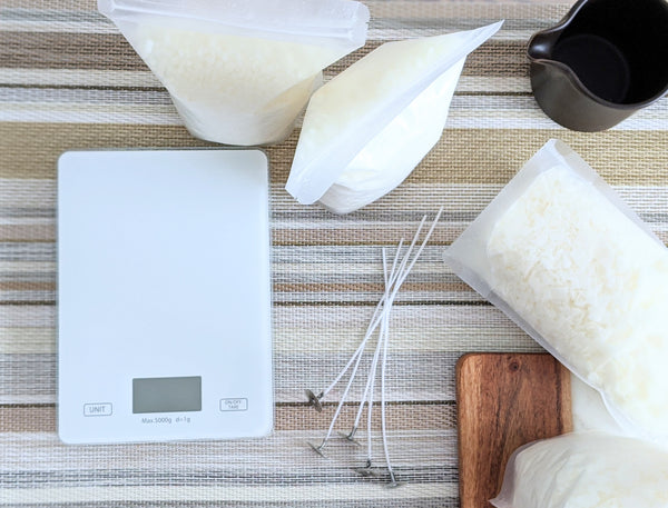 a stylized tabletop with a white scale, wax, wicks, and a pouring pitcher for making candles