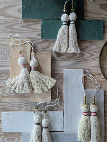 Eight decorative tassels made of off-white cotton thread, each featuring a loop for hanging. The tassels are adorned with various accents including gold-toned metal beads, clear glass beads, and colored thread wraps in pink, green, and red.