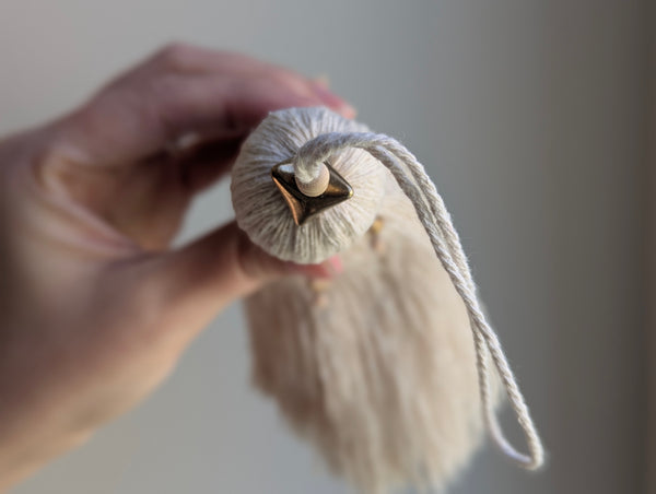 A close-up of a decorative tassel with cream-colored thread, a brown wrapped detail, and brass and wooden beads shown against a neutral gray background