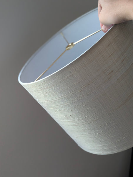 A close-up of a hand holding a drum lampshade made of textured gray silk, showing the white interior and a brass spider washer