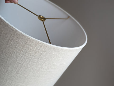 lamp shade made of soft white linen fabric and brass hardware is held at an angle