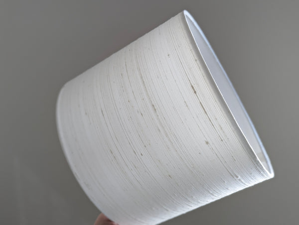 A white drum lampshade with a subtle horizontal textured silk fabric, held by a hand against a plain gray background.