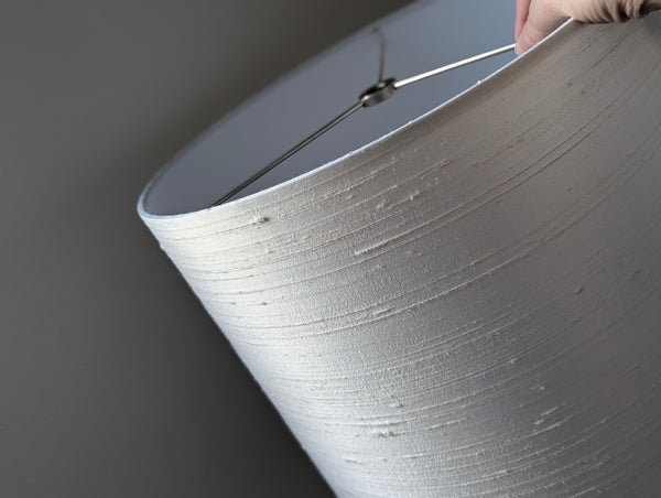 Close-up of a white textured silk lampshade against a dark gray background. A small portion of a hand is visible at the top edge holding the shade.