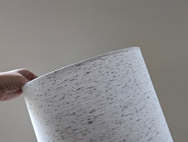 close up of a hand holding a white lamp shade with small black flecks throughout the fabric resembling an oreo blizzard