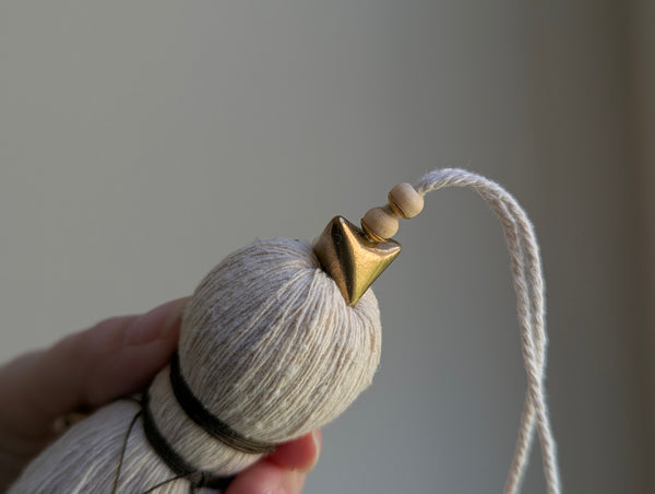 A close-up of a decorative tassel with cream-colored thread, a brown wrapped detail, and brass and wooden beads shown against a neutral gray background