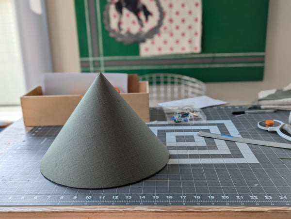 olive green linen fabric lamp shade in cone shape sitting on a cutting mat on a work table with rulers and other tools scattered around it