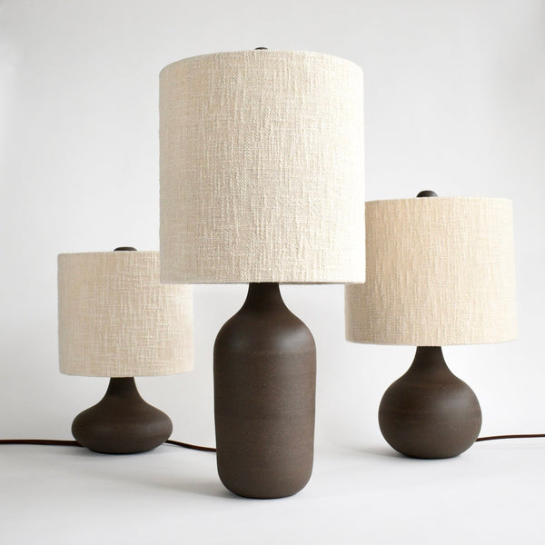 A collection of three CG Ceramics Small Table Lamps with the tallest style in the middle, a round, gumball version to the right and a genie bottle shape to the left. Each of the three lamps is in peat glaze with handmade boucle lamp shades.
