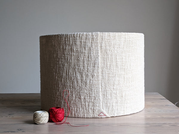 a cream colored, textured fabric lamp shade with red triangle stitch detail sits on a light wood table next to two spools of thread