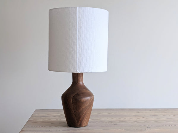 On top of a light wood table, a walnut table lamp sits showing the back seam of the white crosshatch lamp shade that features the triangle stitch detail