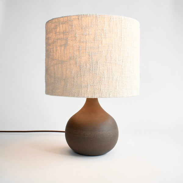 A round, dark brown ceramic table lamp is turned on to show off the handmade boucle lamp shade in cream