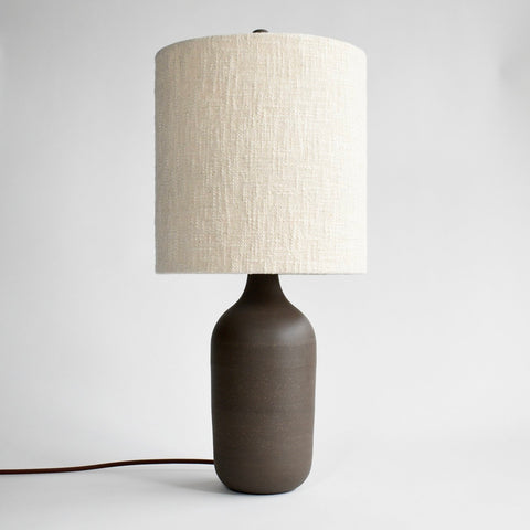 A handmade, deep brown ceramic table lamp with a brown cord and a cream colored boucle lamp shade
