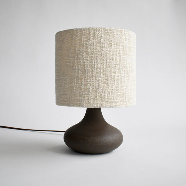 A curvy, dark brown ceramic table lamp is turned on to show off the handmade boucle lamp shade in cream