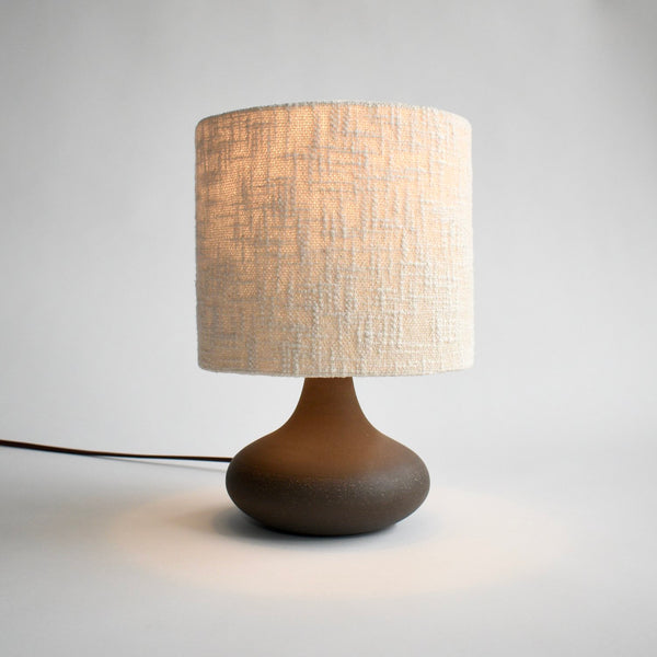 A curvy, dark brown ceramic table lamp is turned on to show off the handmade boucle lamp shade in cream