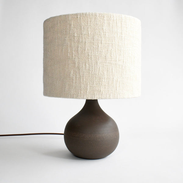 A handmade, deep brown ceramic table lamp with a brown cord and a cream colored boucle lamp shade