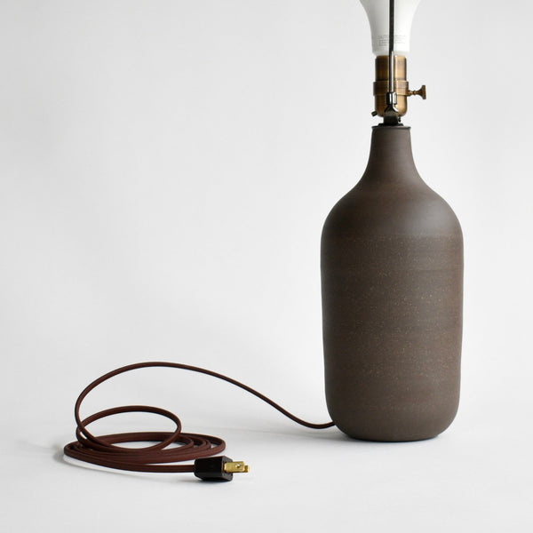 A dark brown cord and plug are attached to a small ceramic table lamp in a dark brown glaze. The shade has been removed showing off the beautiful brown glazed lamp base.