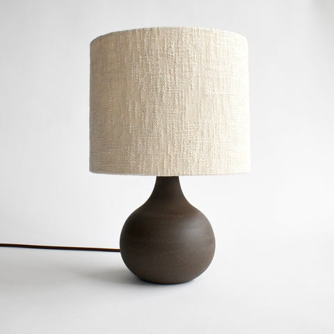 A handmade, deep brown ceramic table lamp with a brown cord and a cream colored boucle lamp shade