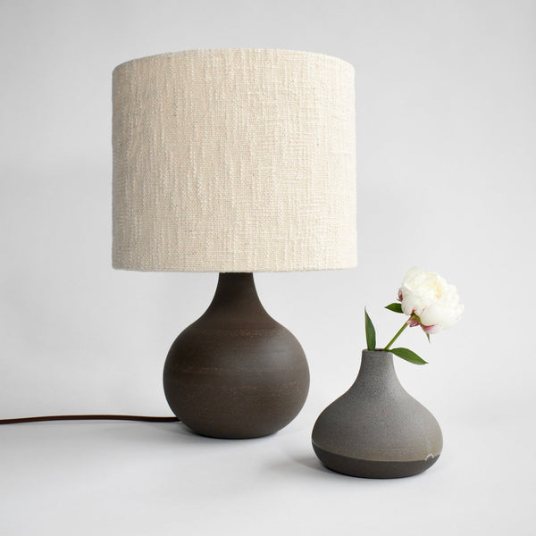 A handmade, deep brown ceramic table lamp with a brown cord and a cream colored boucle lamp shade sits next to a grey glazed vase of a similar shape.