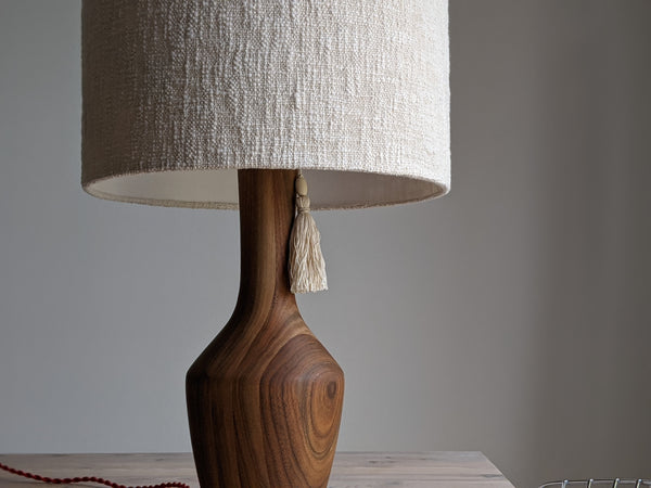 a walnut table lamp with a cream boucle lamp shade and a simple cream colored tassel sits on a light wood table